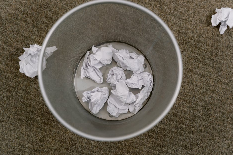 A cylindrical metal bin with a smooth, matte finish, positioned on a brown, textured carpeted floor, is partially filled with crumpled white paper waste. Several additional crumpled papers are scatter